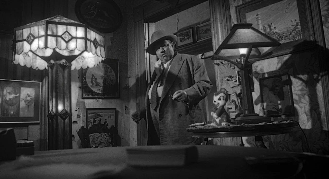 Touch of Evil (1958) movie still: medium — A cluttered room features wallpapered walls with multiple framed pictures and ornate table lamps. A stout…