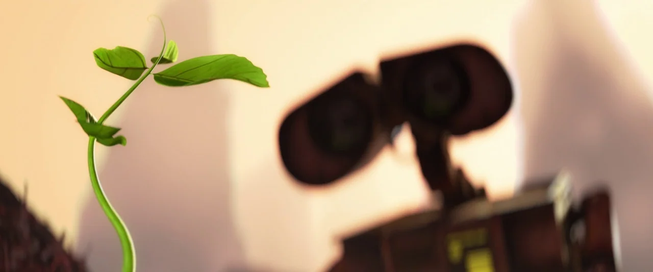 Wall-E (2008) movie still: closeup — A small green plant grows out of dry dirt in a desolate landscape. A robotic character is visible in the…