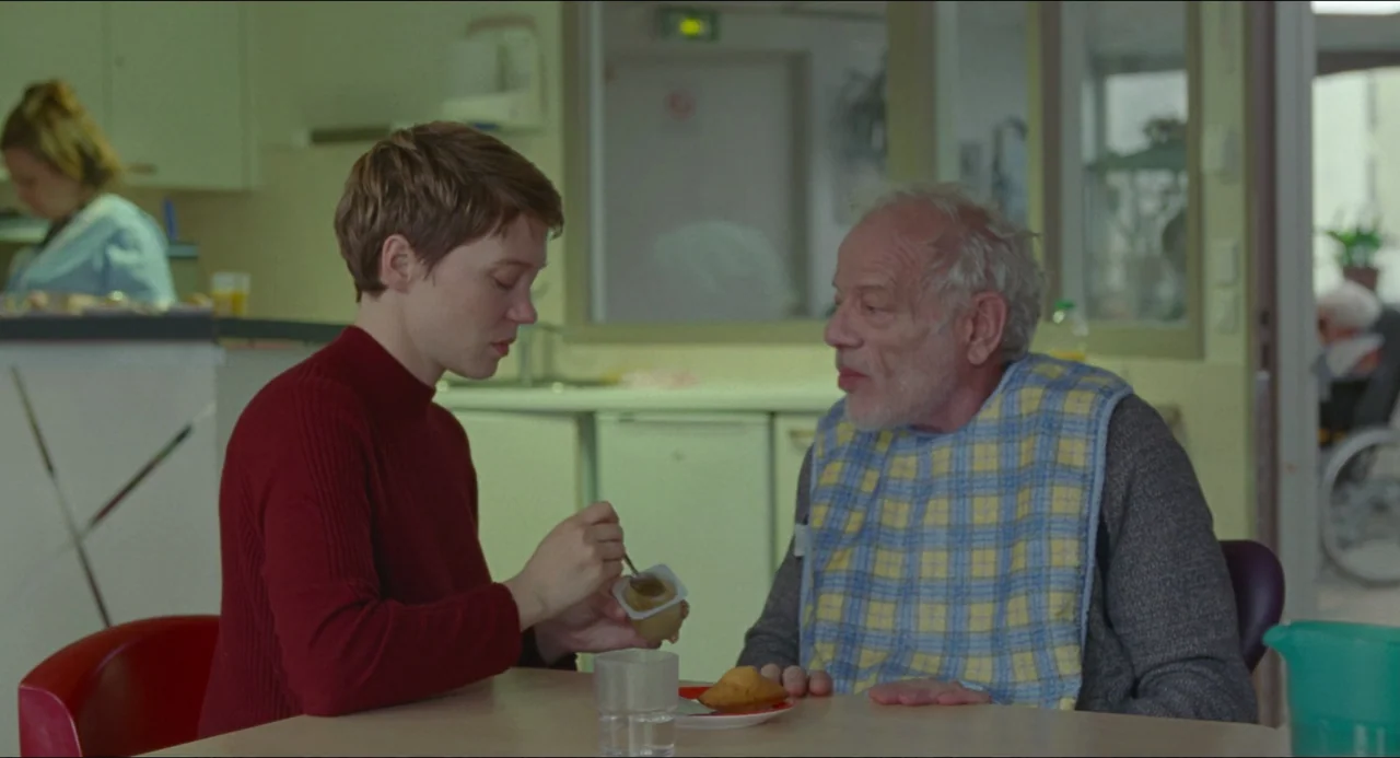 One Fine Morning (2022) movie still: medium — A well-lit medical facility with pale walls and institutional furniture. A young woman sitting at a…