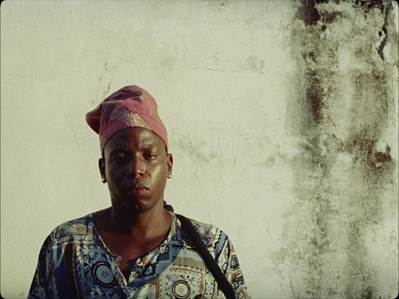 Guava Island (2019) movie still: medium — A man stands in front of a textured, off-white exterior wall with prominent vertical staining. He is…