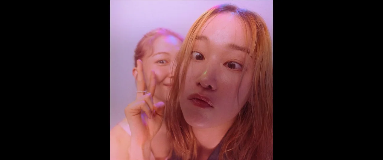 Ballerina (2023) movie still: closeup — Two young women are positioned inside a compact, brightly lit space. One woman stands slightly behind the…