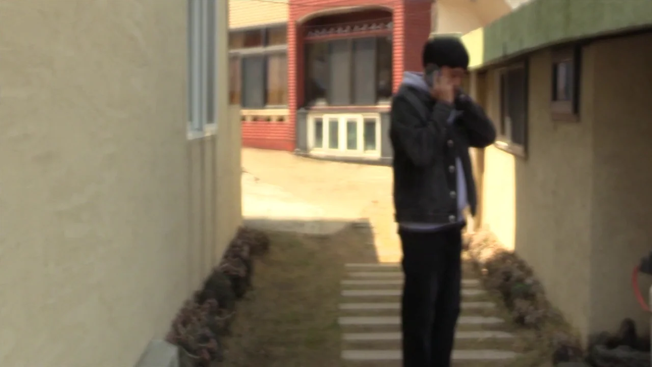 In Water (2023) movie still: fullbody — A narrow residential passage is flanked by beige stucco walls and low landscaping. A young man walks toward…