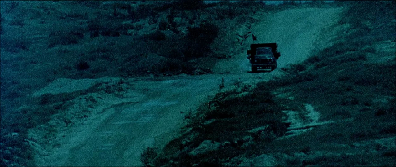 Black Hawk Down (2001) movie still: wide — An unpaved, dusty track winds through a dark, rolling hillside terrain at night. A lone dark truck with…