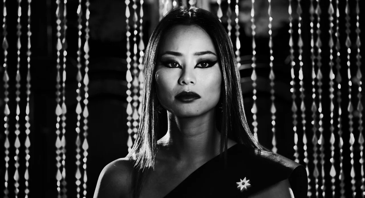 Sin City: A Dame to Kill For (2014) movie still: closeup — A woman stands in front of a curtain made of reflective hanging beads. She maintains a…