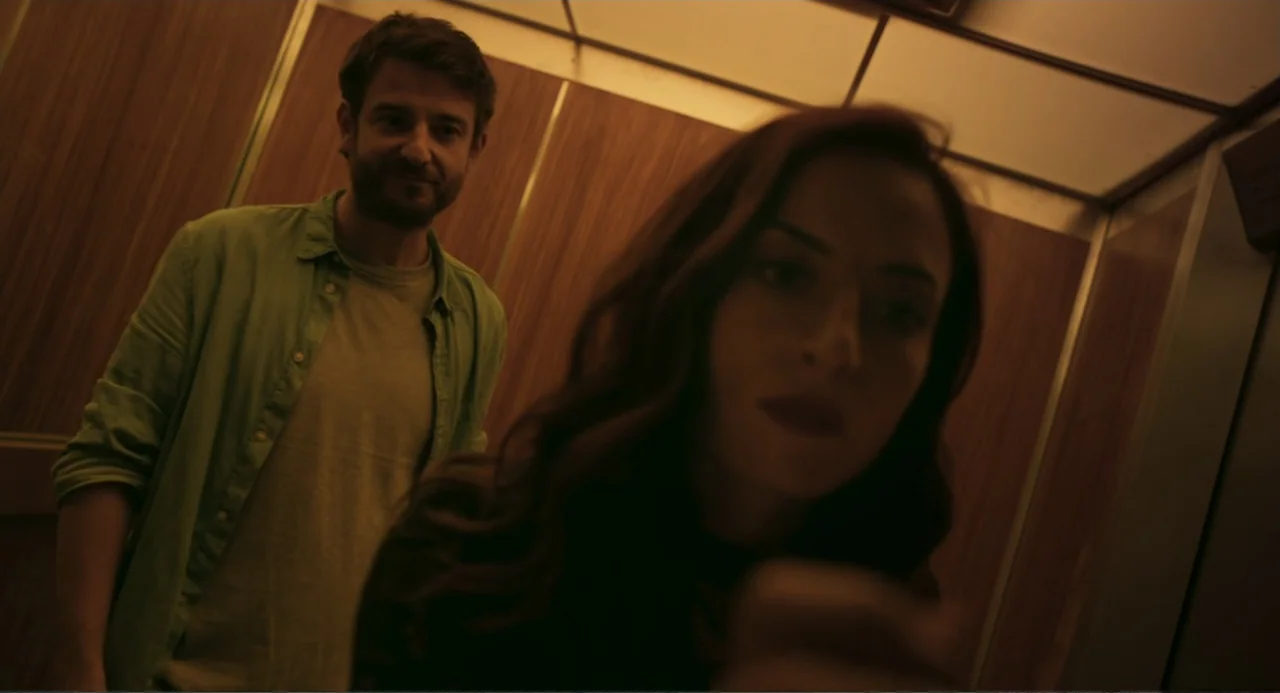 The Lift (2021) movie still: medium — A confined elevator car with wood-paneled walls and a ceiling light panel. A woman in the foreground reaches…