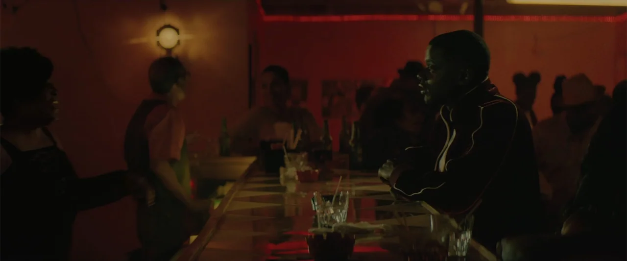 Queen & Slim (2019) movie still: medium — A dimly lit bar glows with intense red neon light reflecting on surfaces. A man sits at the wooden bar…