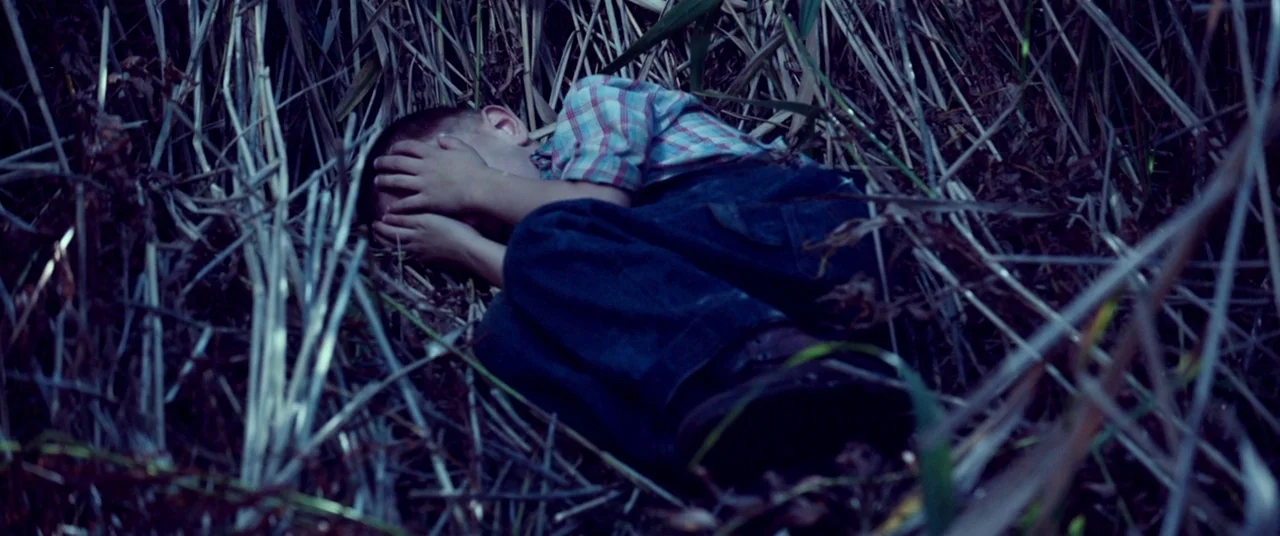 The House That Jack Built (2018) movie still: medium — A child is curled up tightly amidst dense, dry reeds in the dark night. The child hides their…