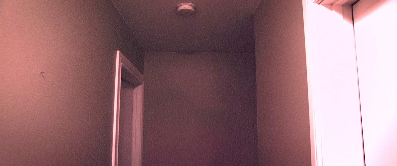 Skinamarink (2022) movie still: establishing — A residential hallway features plain walls and a single ceiling-mounted smoke detector. No occupants…