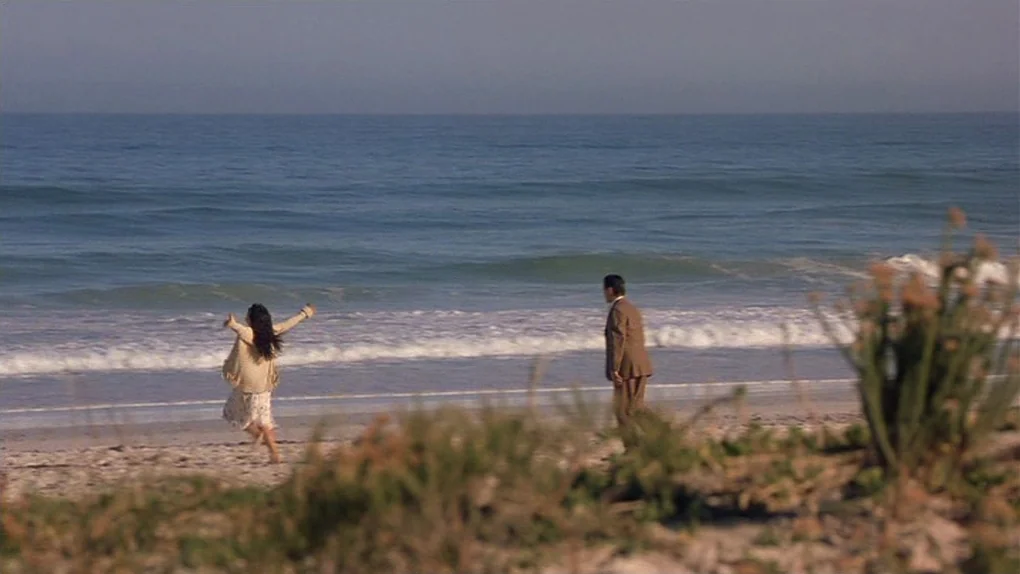 Ask The Dust (2006) movie still: wide — A wide sandy beach stretches along the shoreline with gentle ocean waves rolling in. A woman in a light…