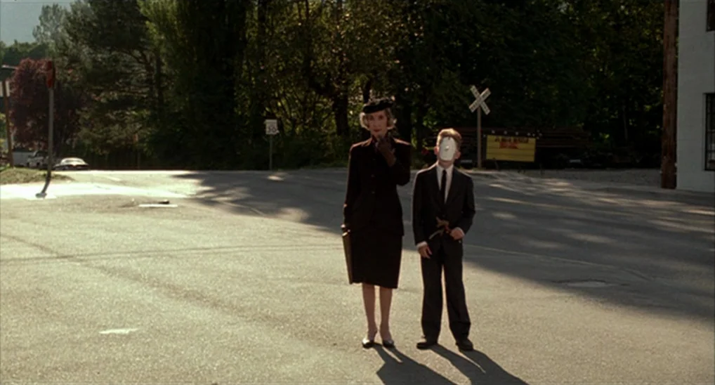 Twin Peaks: Fire Walk With Me (1992) movie still: fullbody — An asphalt intersection is bordered by dark trees and a white wall in daylight. A woman…