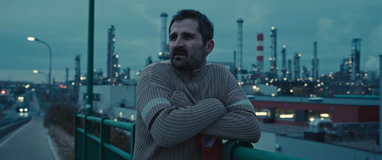 R.M.N. (2022) movie still: medium — An industrial facility with illuminated smokestacks stands behind a metal railing at dusk. A man in a beige…