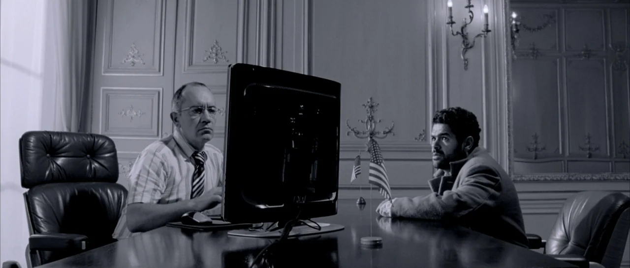 Angel-A (2005) movie still: medium — A formal office space features paneled walls and a large, dark-wood desk. Two men sit opposite each other with…