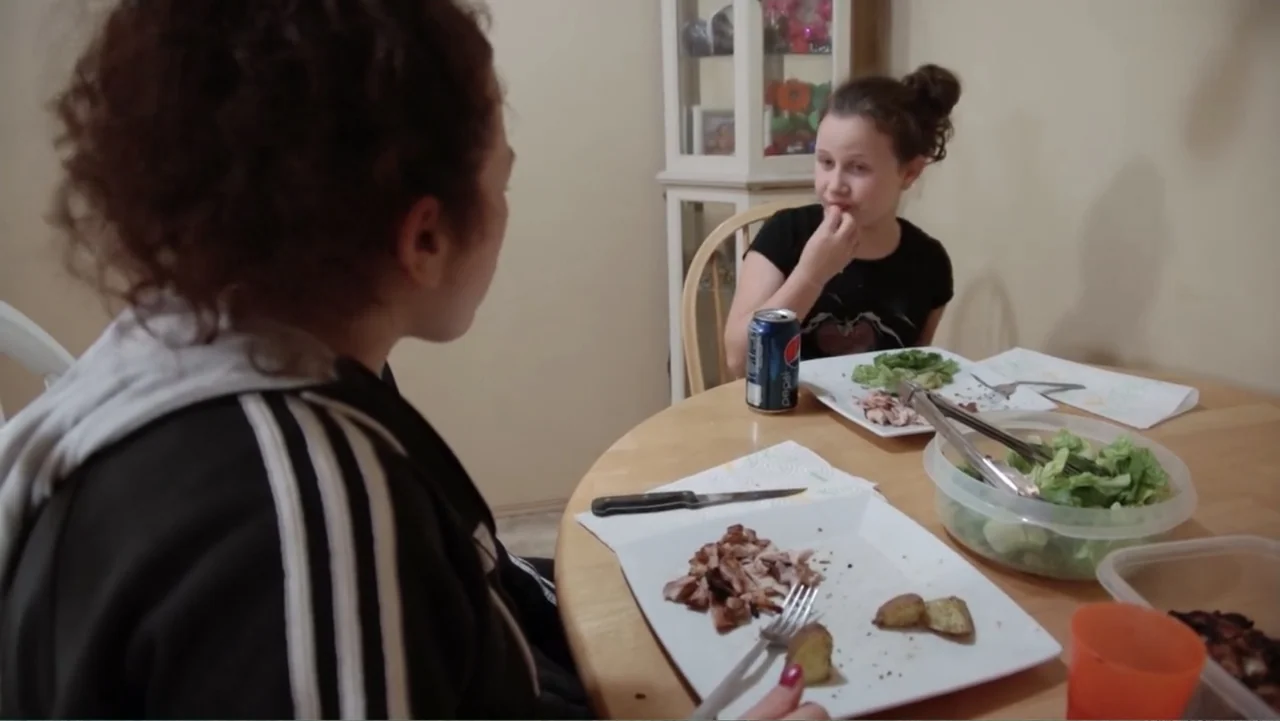 A Dangerous Son (2018) movie still: medium — A dining table sits in a room with a white china cabinet behind. Two people are eating grilled meat and…