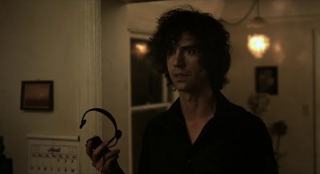 The Future (2011) movie still: medium — A dimly lit residential house interior with a calendar on the wall. A young man with dark curly hair holds a…