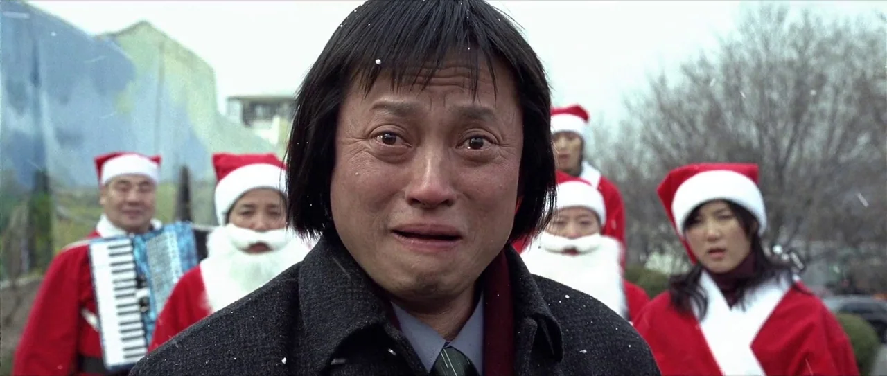 Lady Vengeance (2005) movie still: closeup — A man stands in the foreground with a group of people wearing Santa hats behind. The background…