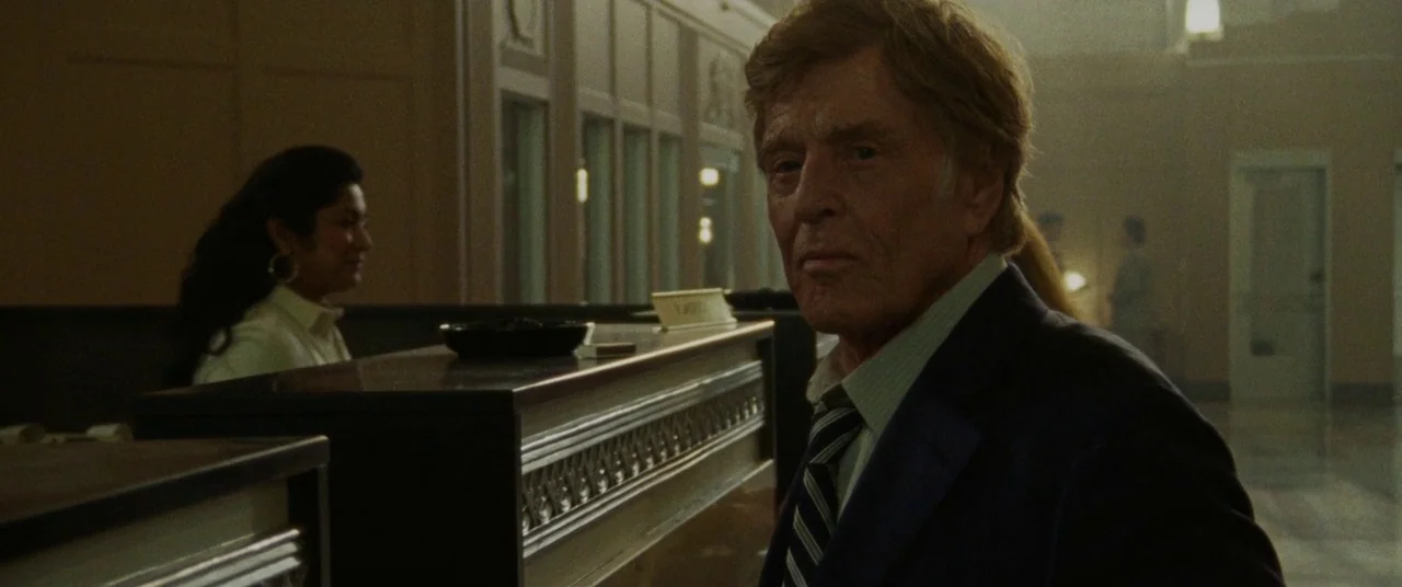 The Old Man & The Gun (2018) movie still: medium — A quiet bank lobby with dark wooden counters and pale, institutional wall paneling. An older man…