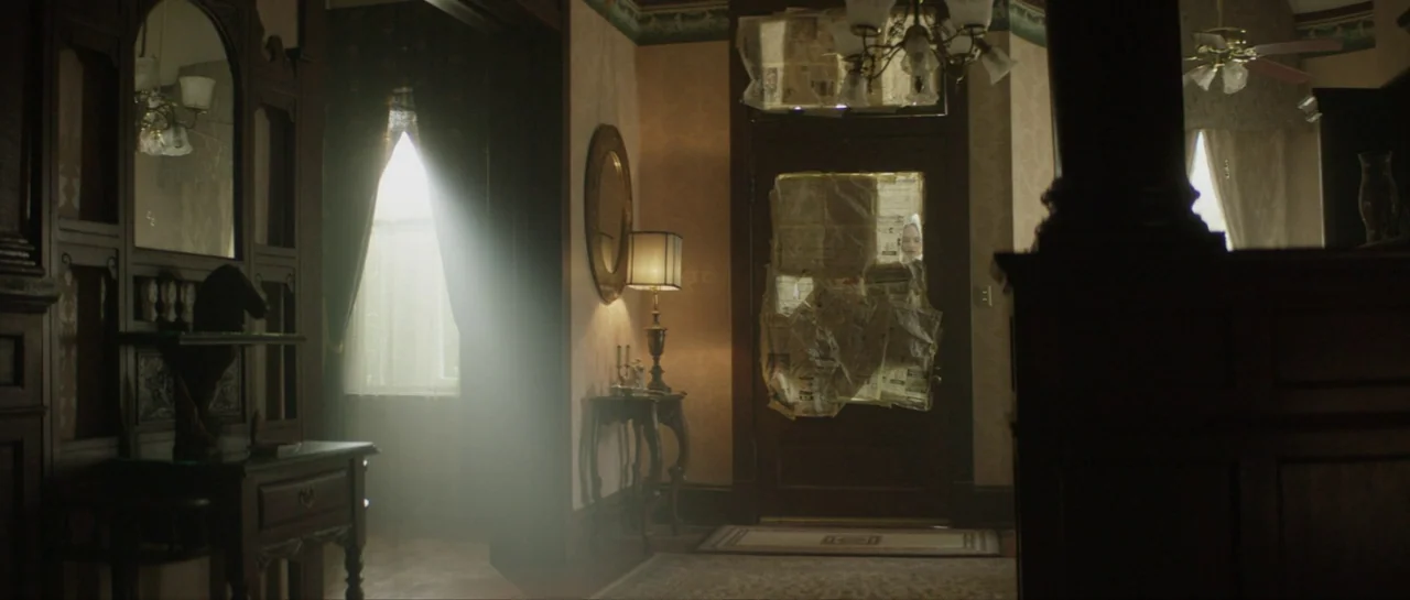 Suitable Flesh (2023) movie still: wide — A dimly lit entryway with wood furniture and patterned wallpaper features a door covered in newspaper.…