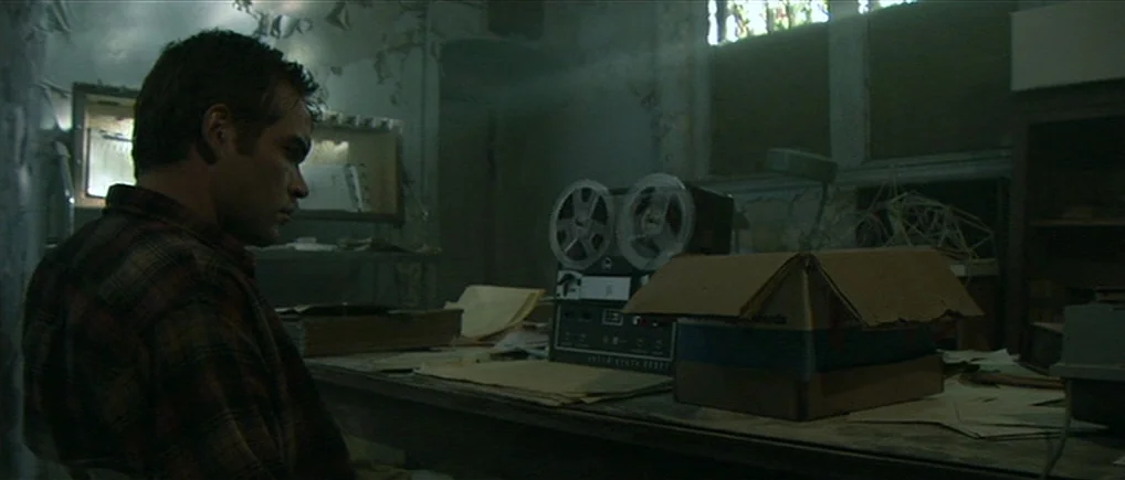 Session 9 (2001) movie still: medium — A cluttered room with peeling wallpaper and scattered papers on a work desk. A man sits in profile watching a…