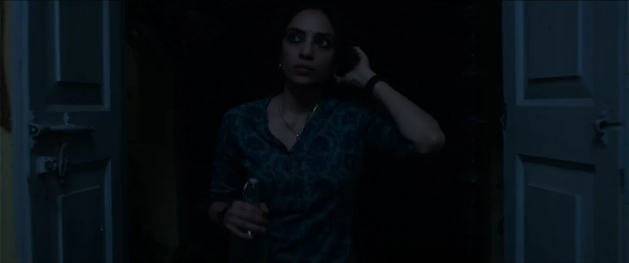 Cheekatilo (2026) movie still: medium — A woman stands in a dark doorway framed by wooden panels at night. She holds a glass bottle while reaching…