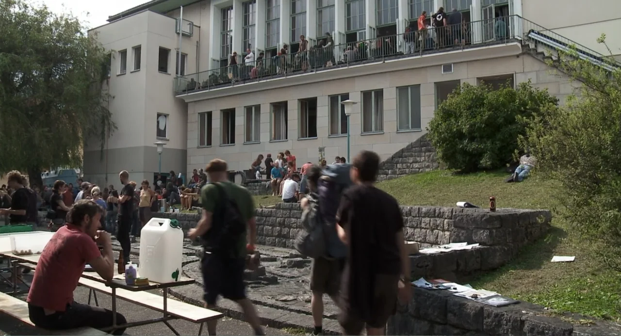 Az X- Kryptonit projekt (2020) movie still: wide — A stone courtyard sits before a large institutional building with a second-story balcony. Many…