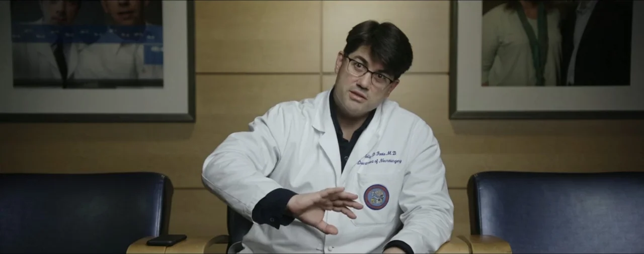 Hunting for Hedonia (2019) movie still: medium — A man sits in an office waiting area with wood paneled walls and dark chairs. A male medical…