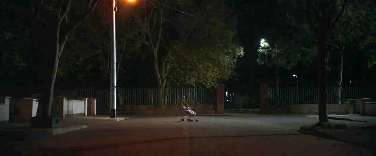 You Are Not My Mother (2021) movie still: wide — An empty road at night is surrounded by tall trees and a gated fence. A single baby stroller stands…