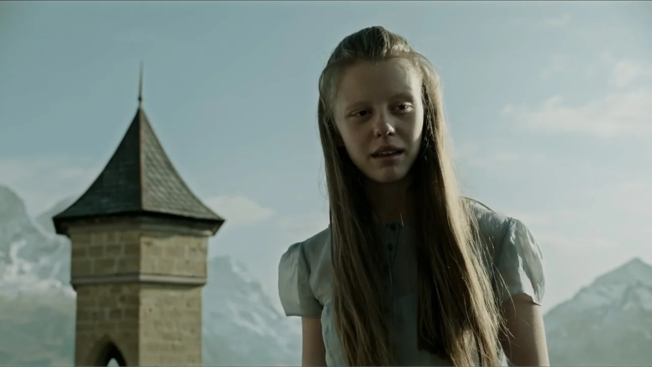 A Cure for Wellness (2017) movie still: medium — A pale teenage girl stands before a weathered stone tower in a mountainous landscape. The…