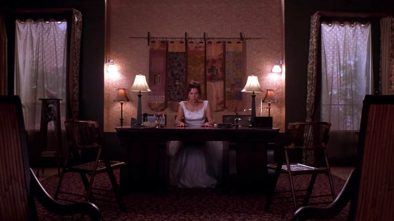 Secretary movie still: medium — A symmetrical room with textured wallpaper and hanging fabric panels features a centered desk. A woman sits at the…