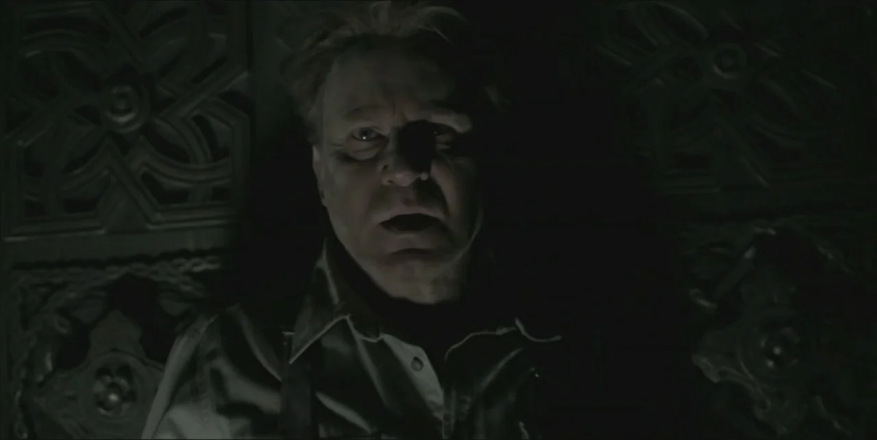 Exorcist: The Beginning (2004) movie still: closeup — A dimly lit indoor space features a dark wall with ornate carved wood patterns. A man stands…