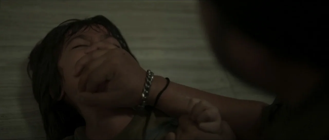 Innocent Vengeance (2023) movie still: closeup — A dimly lit indoor space with a plain, light-colored background. A large hand covers the mouth of a…