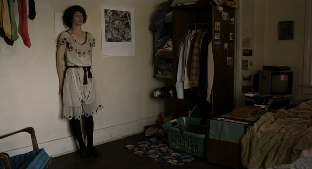 The Future (2011) movie still: fullbody — A cluttered room features off-white walls, an open wardrobe, and an old television set. A woman in a dress…