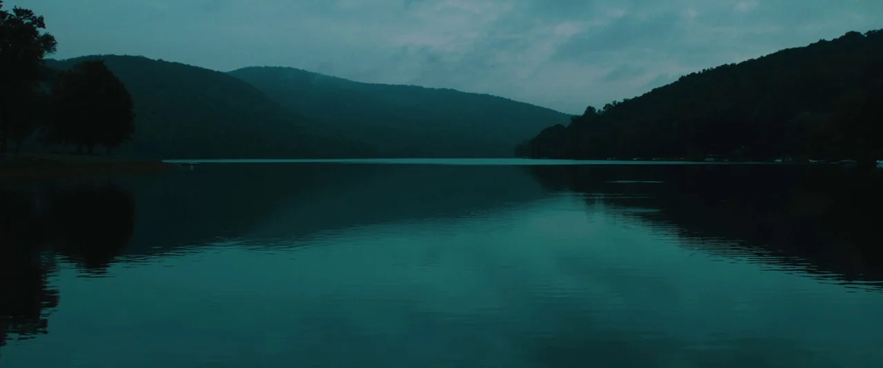 Kiss of the Damned (2012) movie still: establishing — A dark, calm lake is surrounded by densely forested hills under a cloudy sky. The water…