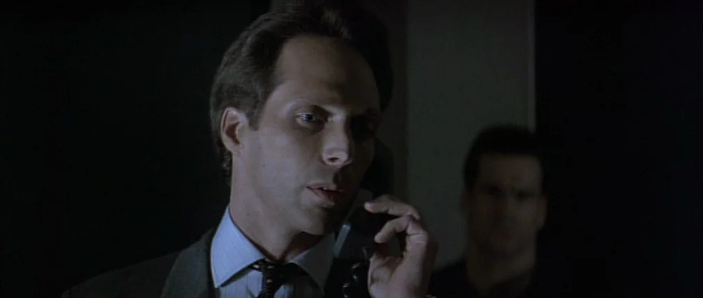 Heat (1995) movie still: closeup — A dark indoor setting with limited illumination on the subjects. The primary man is speaking on a telephone in…
