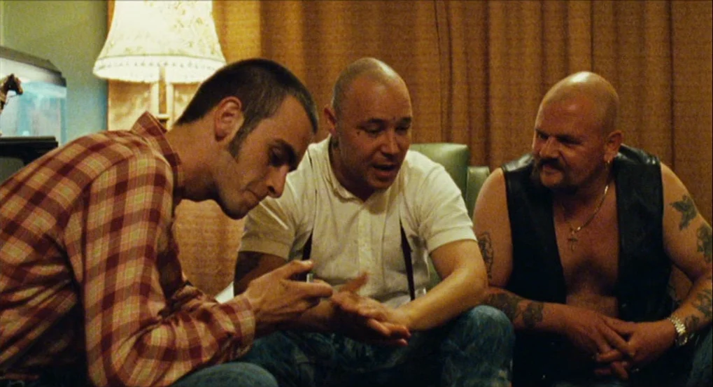 This Is England (2006) movie still: medium — A dimly lit room with brown curtains and a fringed lampshade. Three men sit closely together engaged in…