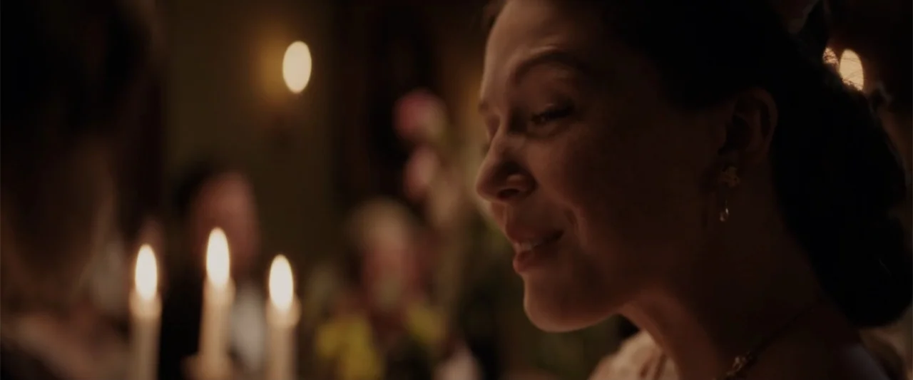 I Heard the Bells (2022) movie still: closeup — A dimly lit room features a table set with lit candles and floral arrangements. A woman in period…
