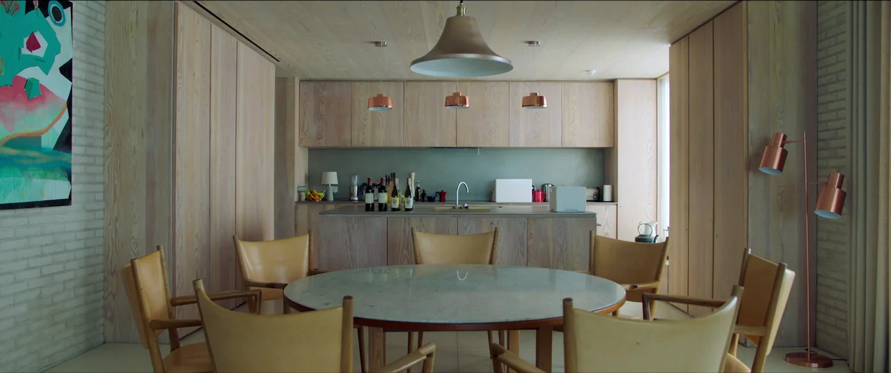 The Feast (2021) movie still: wide — A modern dining area features light wood cabinetry and a marble-topped circular table. Several leather…