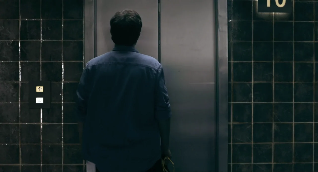 The Lift (2021) movie still: medium — A tiled interior lobby features dark wall tiles and metallic elevator doors. A man in a blue shirt stands…