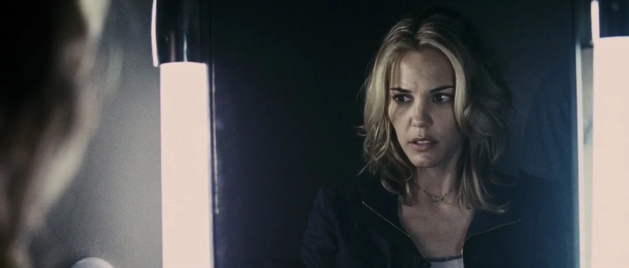 The Midnight Meat Train (2008) movie still: closeup — A dimly lit room features a woman standing before a brightly lit vanity mirror. She stares…