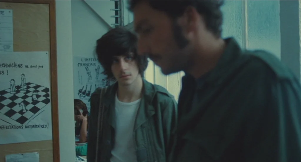 Something in the Air (2012) movie still: medium — A narrow hallway features a bulletin board with posters containing black and white illustrations.…
