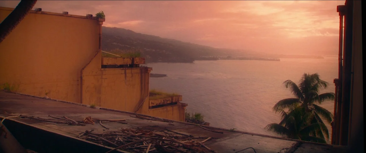 Pacifiction (2022) movie still: establishing — A weathered concrete rooftop overlooking a calm bay during a hazy sunset. Dried twigs and debris are…