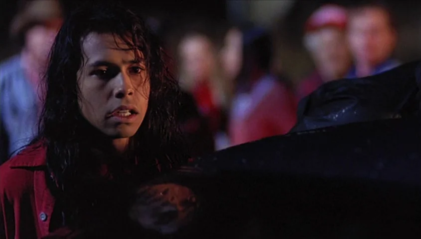 Dance Me Outside (1994) movie still: closeup — An outdoor night scene shows a young man amidst a blurred crowd of onlookers. He looks toward the…