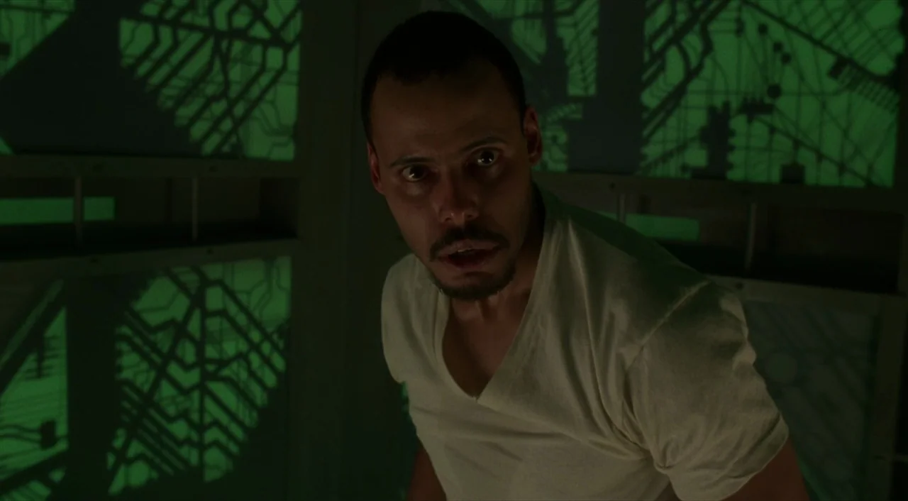 Cube (1997) movie still: medium — A small interior space with translucent panels showing green circuit patterns on the walls. A man stares intensely…