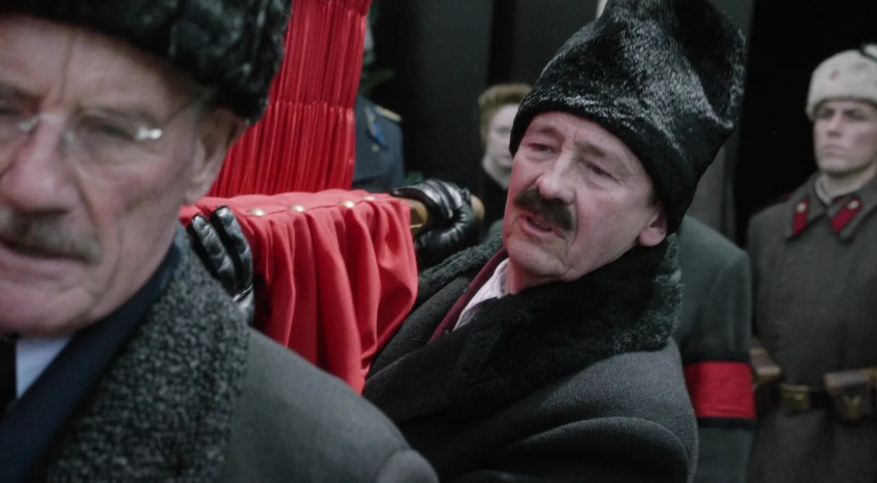 The Death of Stalin (2017) movie still: medium — A group of somber men carry a casket covered in vibrant red cloth indoors. The men wear formal…