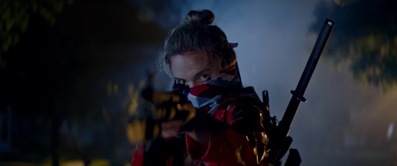 Assassination Nation (2018) movie still: medium — A dark, foggy residential street at night creates an obscured backdrop for the character. A person…