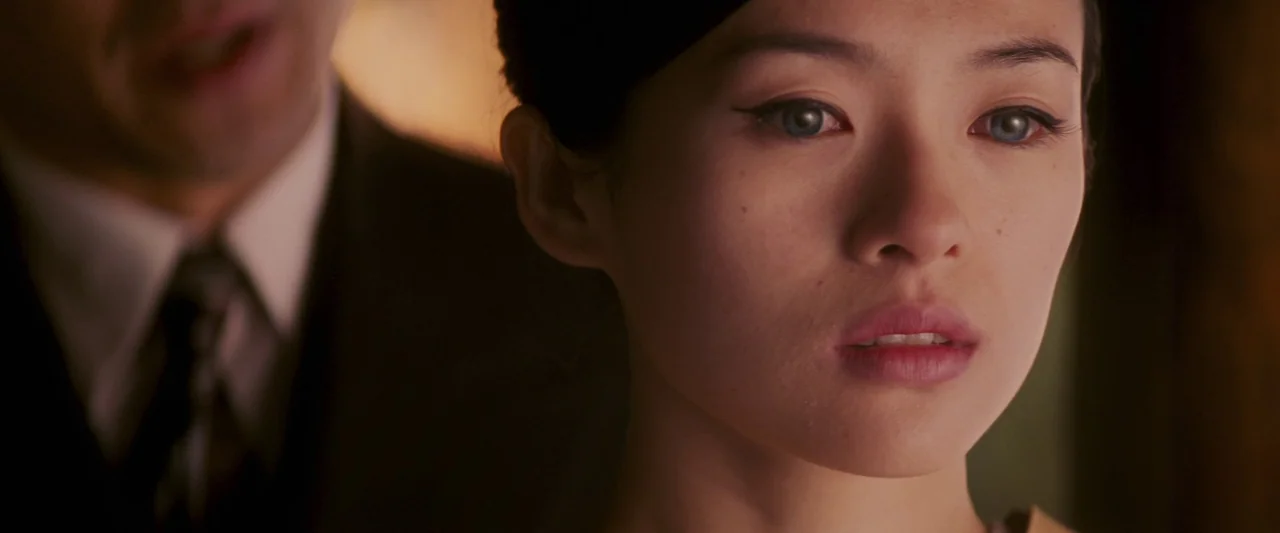 Memoirs of a Geisha (2005) movie still: closeup — A close-up view shows a young woman's face in soft light. A man in a suit is partially visible…