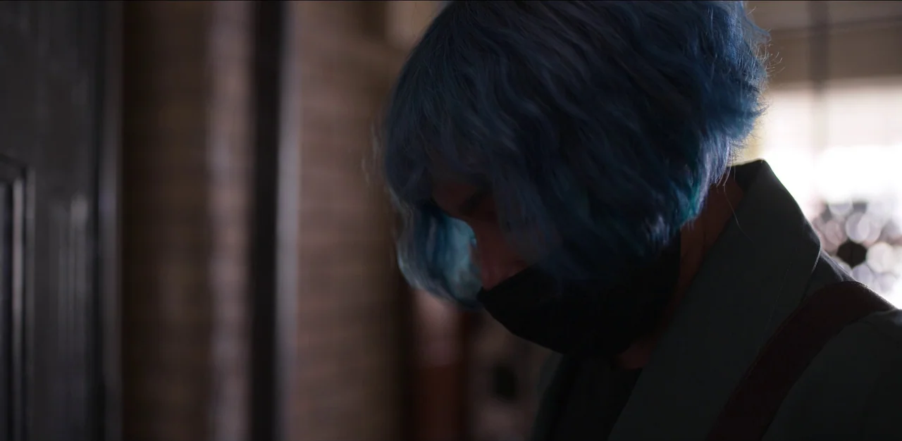 Kimi (2022) movie still: closeup — A person stands in a dimly lit indoor space with brick textured walls. The individual with vibrant blue hair and…