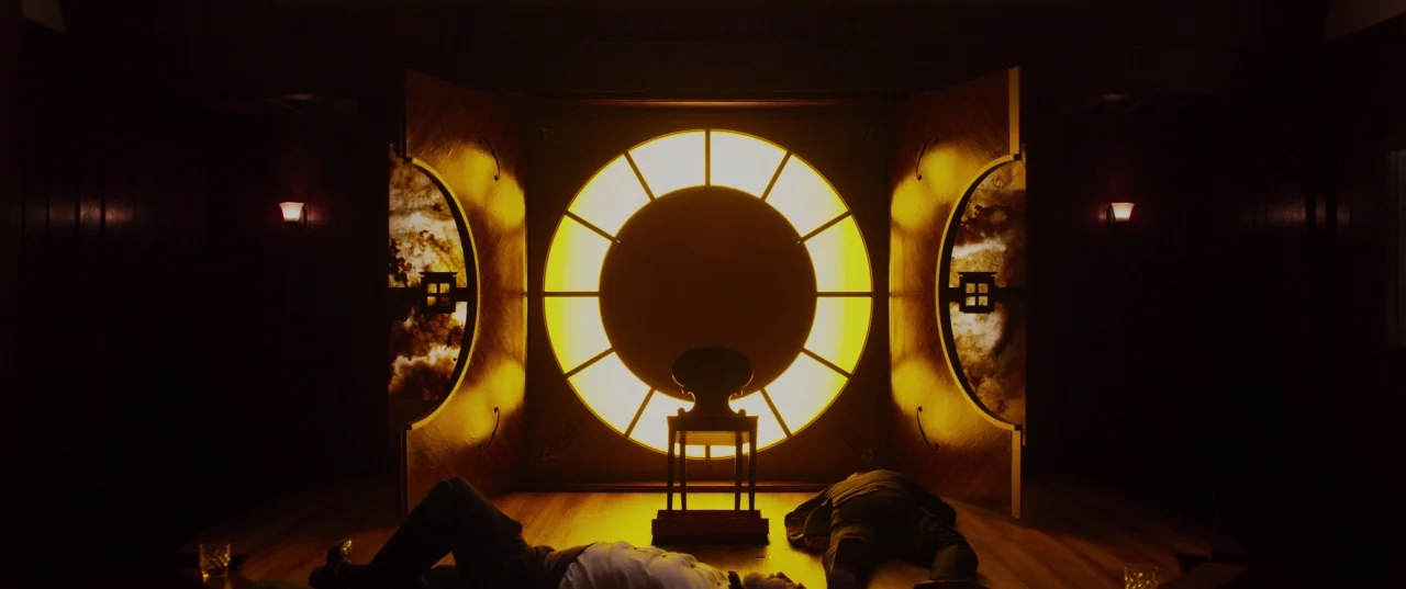 The Perfection (2018) movie still: wide — A dark, dimly lit lounge features a prominent circular screen panel glowing with yellow light. Two men lie…