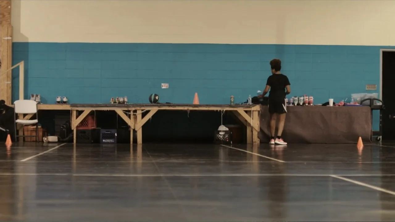 Death of a Child (2017) movie still: wide — A roller skating rink features a blue-painted cinder block wall and a long wooden table. A young person…
