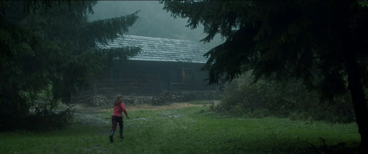 Nightsiren (2022) movie still: wide — A remote cabin with a shingled roof sits in a grassy area amidst forest trees. A small child runs across the…