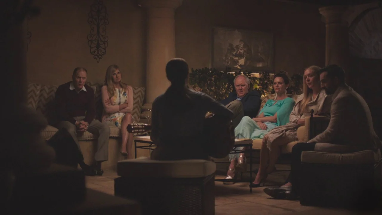 Beatriz at Dinner (2017) movie still: wide — A dimly lit room with stone columns and framed art features an seated group. A person centered in the…